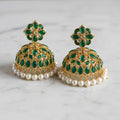 A pair of traditional Indian gold-plated jhumka earrings decorated with green enamel, white pearl beads, and small clear stones, displayed on a light gray marble surface.