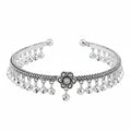 A silver-toned metal cuff bracelet featuring a braided pattern, dangling bell charms, and a central flower motif with a small clear stone, shown on a plain white background.