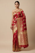 A woman models a red and gold Banarasi silk saree with intricate gold zari motifs and a wide gold border, set against a solid beige studio background.