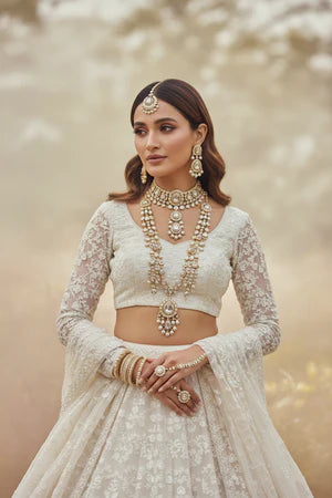 A woman wearing an elegant off-white embroidered traditional outfit with a lace long-sleeve crop top and flared skirt, styled with ornate gold jewelry against a soft beige background.